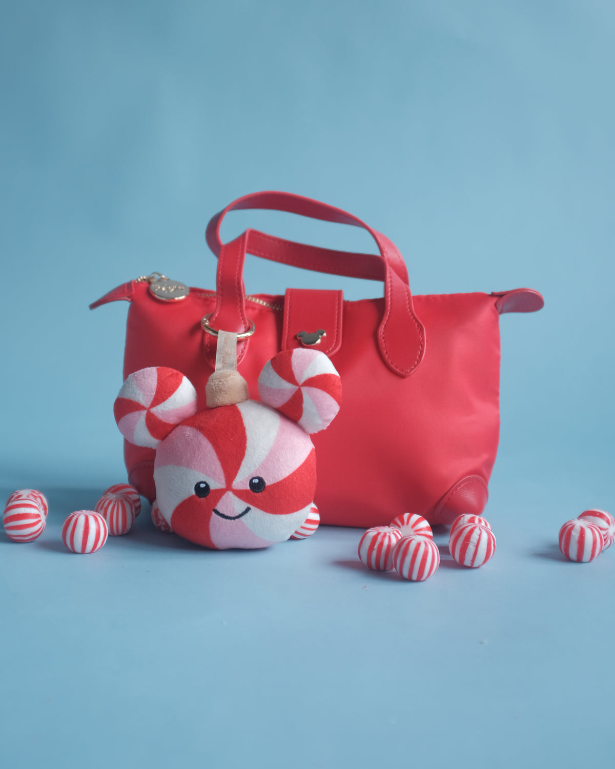 Peppermint Pal bag charm clipped to a red handbag, with peppermint candies on a blue backdrop