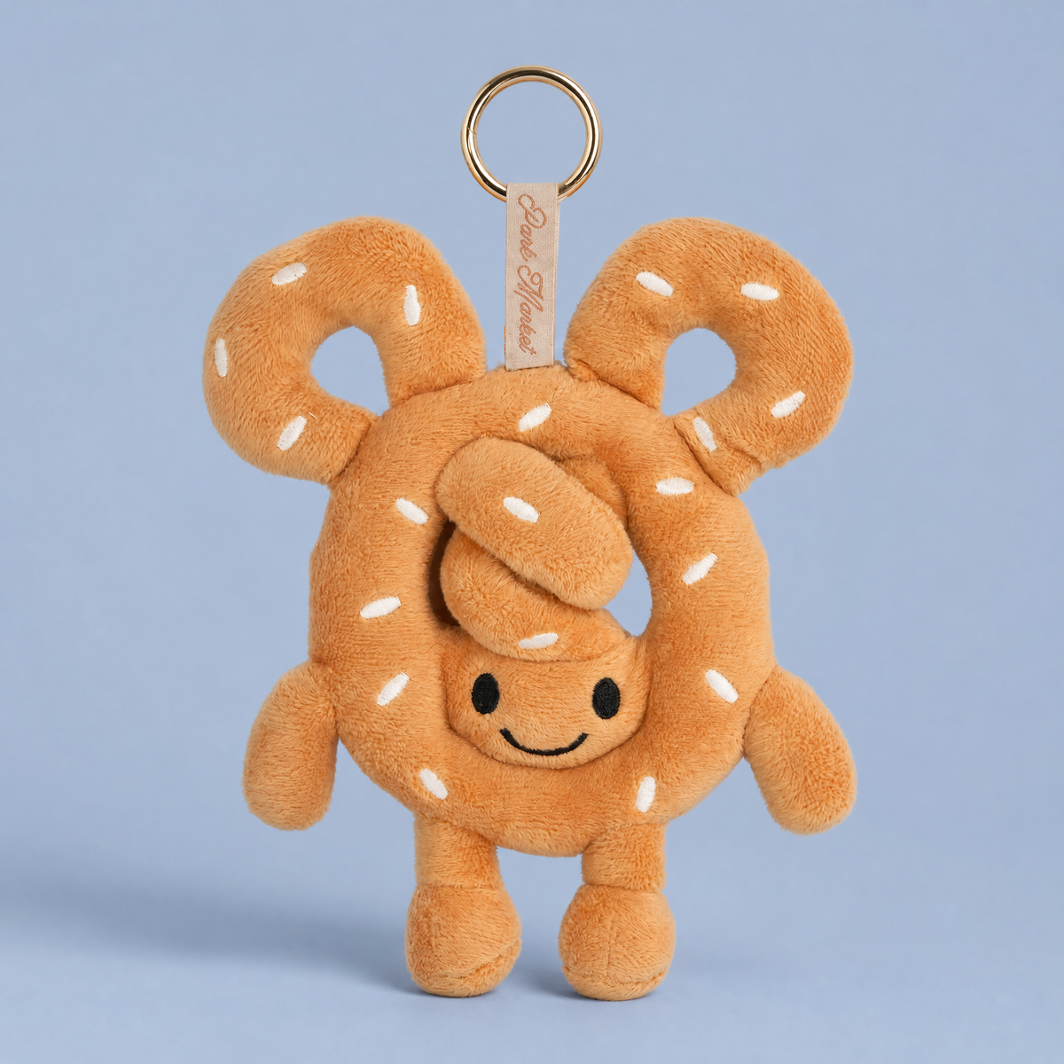 Pretzel plush bag charm with embroidered salt, a smiling face, and little arms and legs, hanging from a gold ring against a light blue background