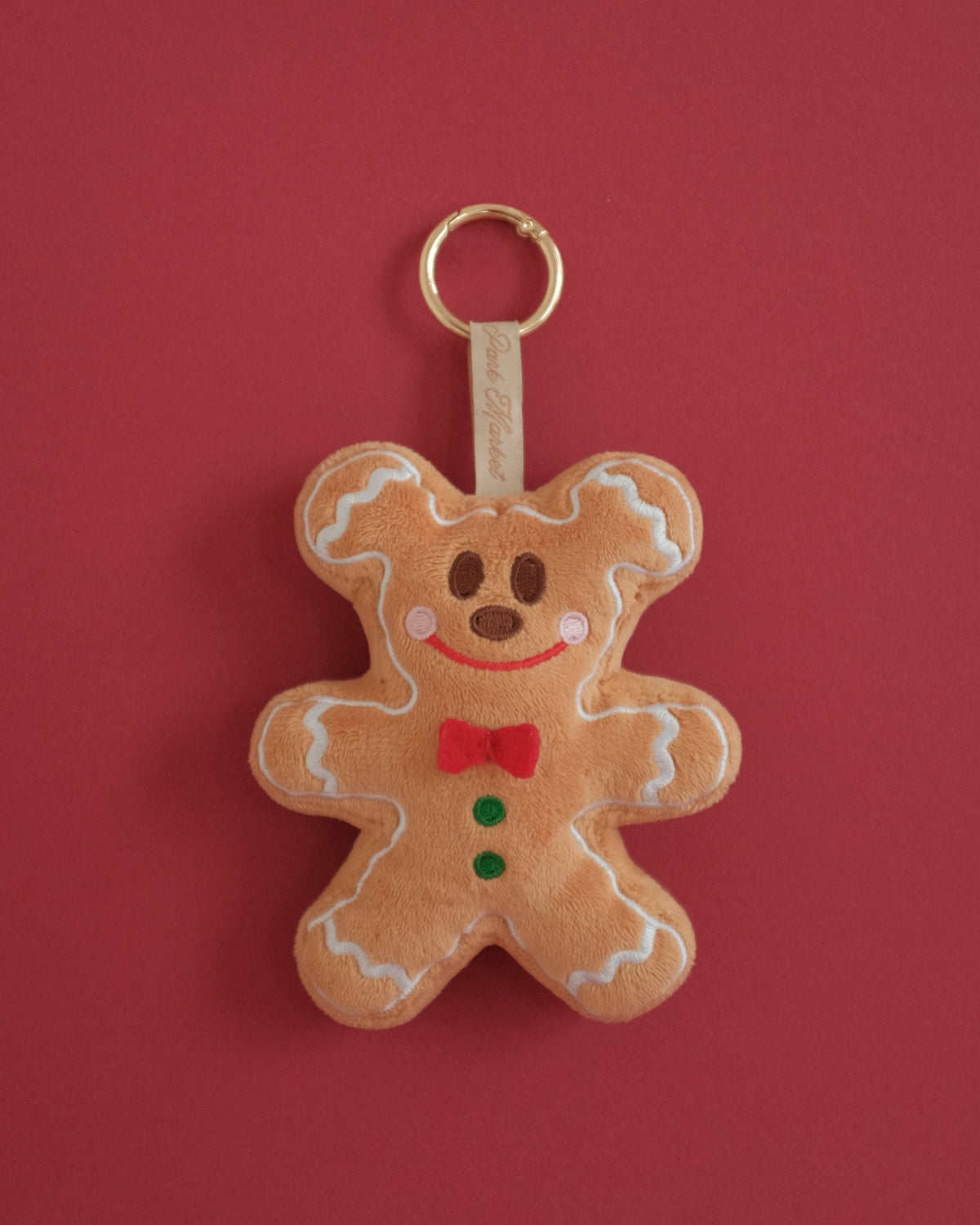 Gingerbread Gent plush bag charm with embroidered frosting, rosy cheeks and a red fabric bow tie, on a red background