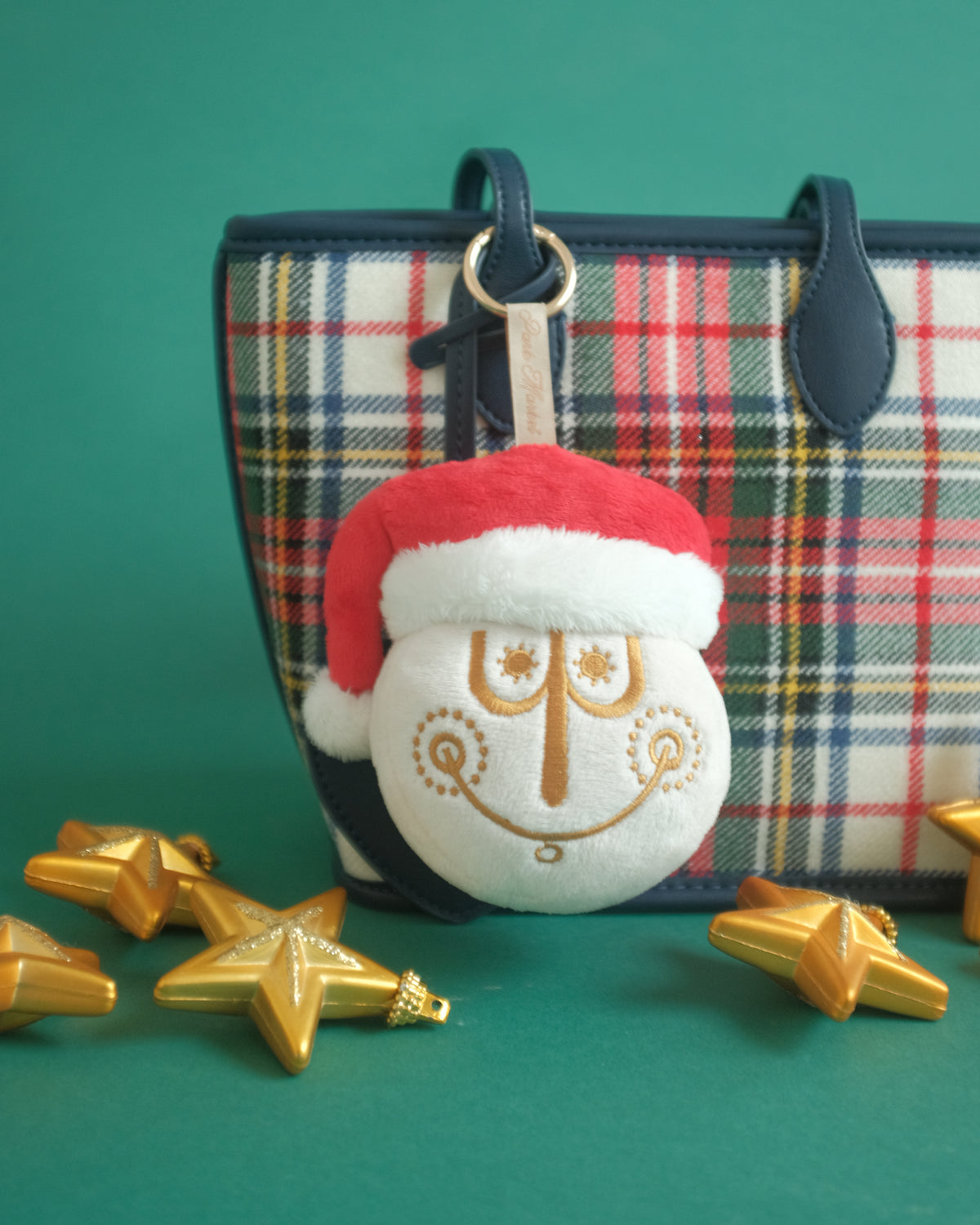Close-up of the Tinsel Time bag charm on a plaid tote, surrounded by gold star ornaments