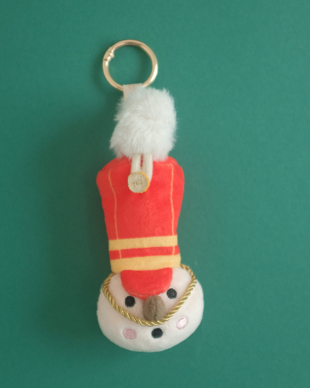 The Merry Marcher plush toy soldier bag charm with a red hat, gold trim and white plume, on a green background