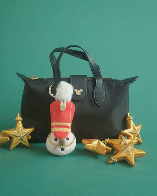 The Merry Marcher bag charm on set: clipped to a black bag, a close-up, gold ornaments and pink gifts
